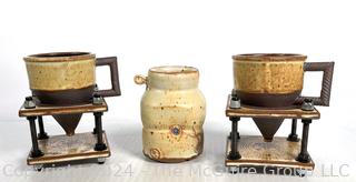 Two artisian coffee/tea mugs with conical bases on stands (perhaps to catch tea leaves) and a third ceramic mug