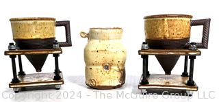 Two artisian coffee/tea mugs with conical bases on stands (perhaps to catch tea leaves) and a third ceramic mug